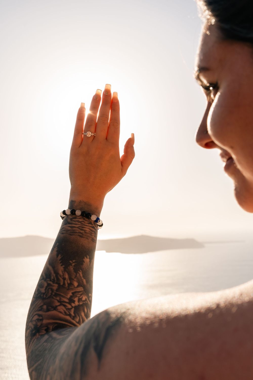 Woman with tattooed arm and ring holds hand up against sunset over ocean in Santorini Greece.