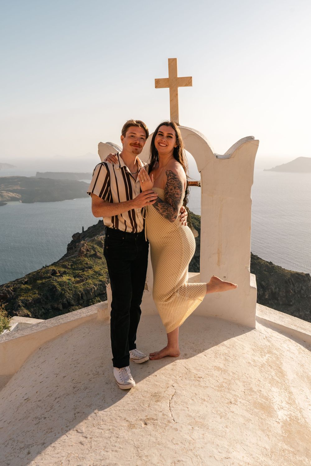 Couple posing by a white cross arch in Santorini, Greece, with stunning caldera views in the background.