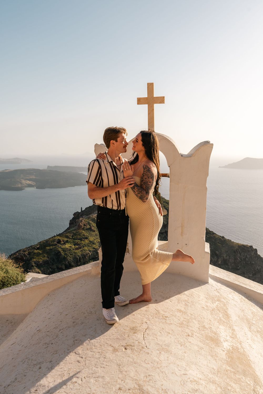 Couple embracing near white cross monument overlooking Santorini caldera at golden hour.