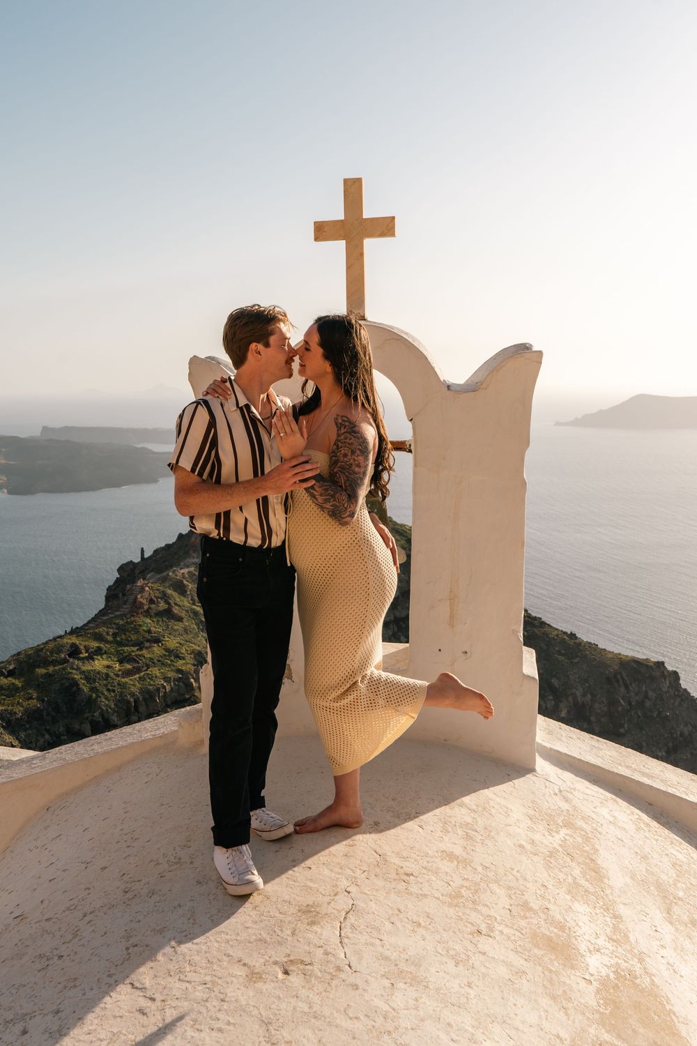 Romantic couple kissing by a white church bell tower with cross in Santorini, Greece, with caldera views.