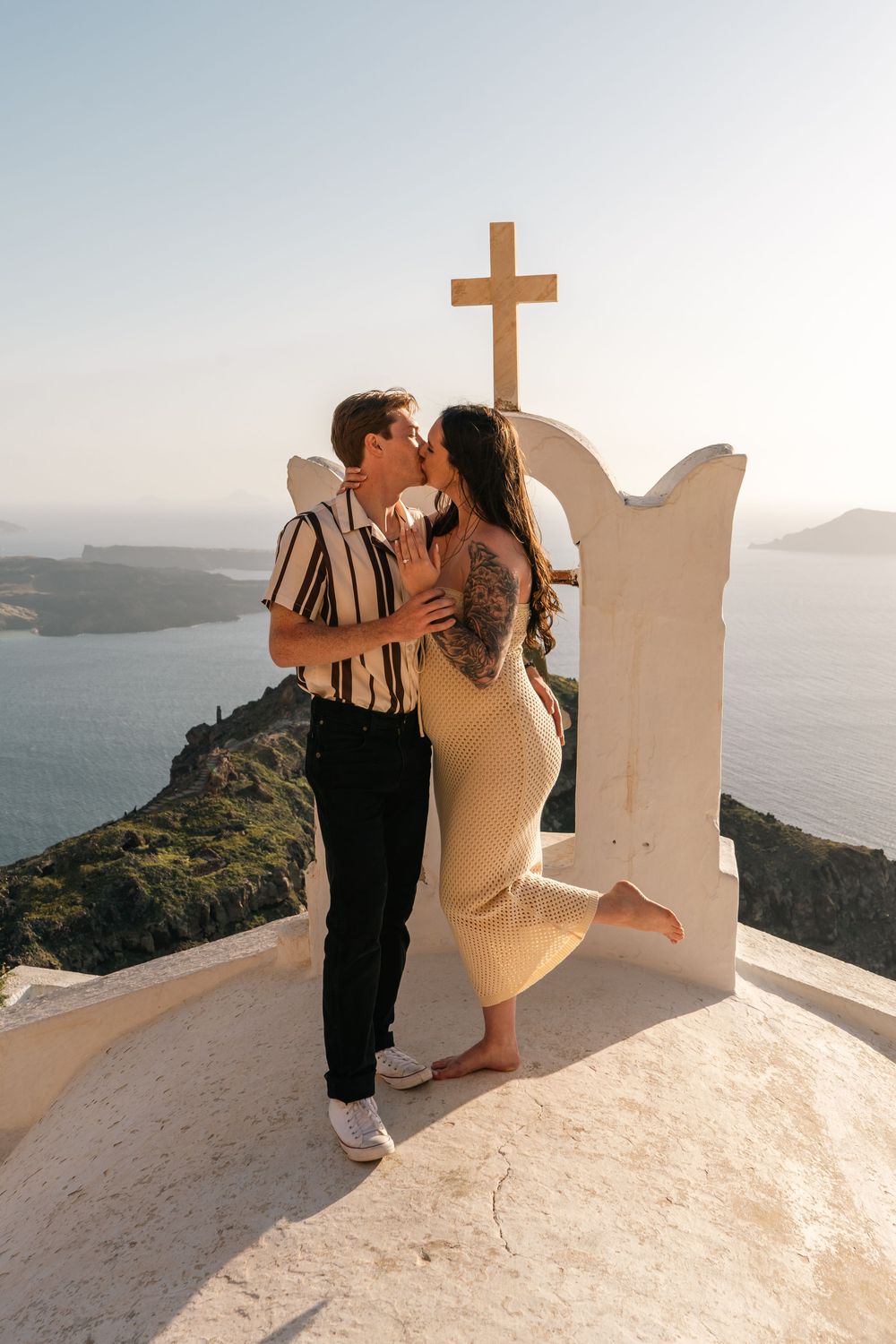 Couple kissing in front of a white cross arch in Santorini, Greece, with caldera views and blue sea in the background.