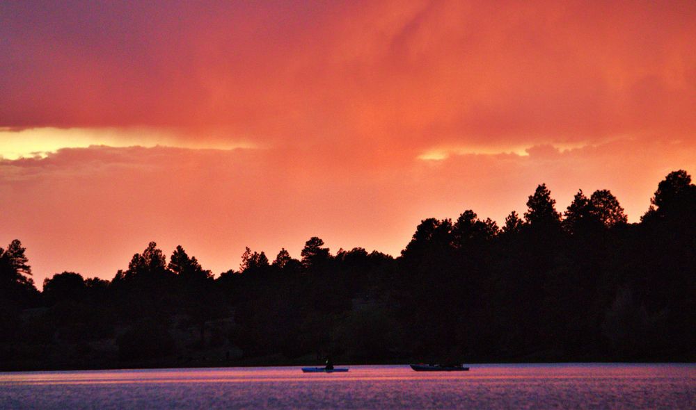 A vibrant orange sunset silhouettes pine forest trees along a lake with boats visible in the purple-tinted water.