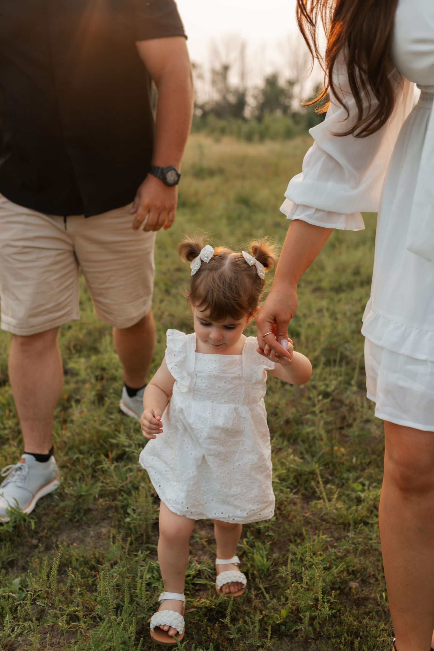 Toddler girl in white dress holding parents hands while walking through green meadow at sunset.