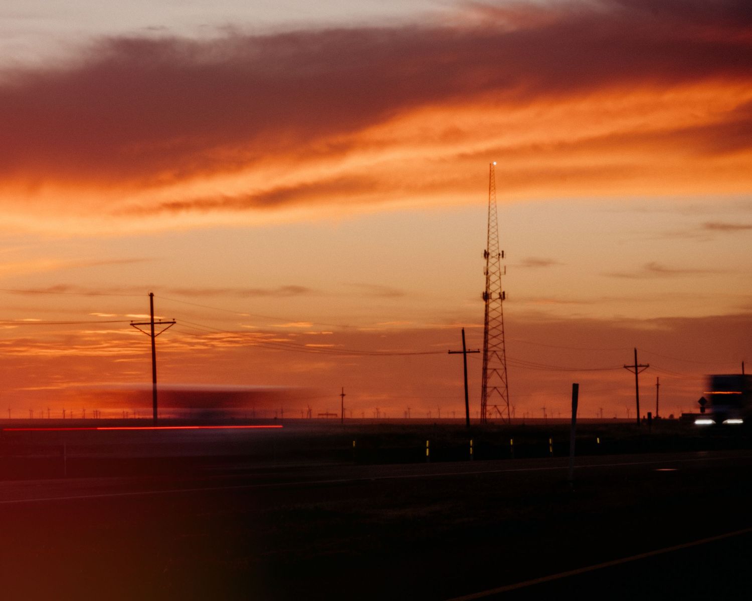 Dramatic orange sunset silhouettes power lines and transmission towers against the sky.