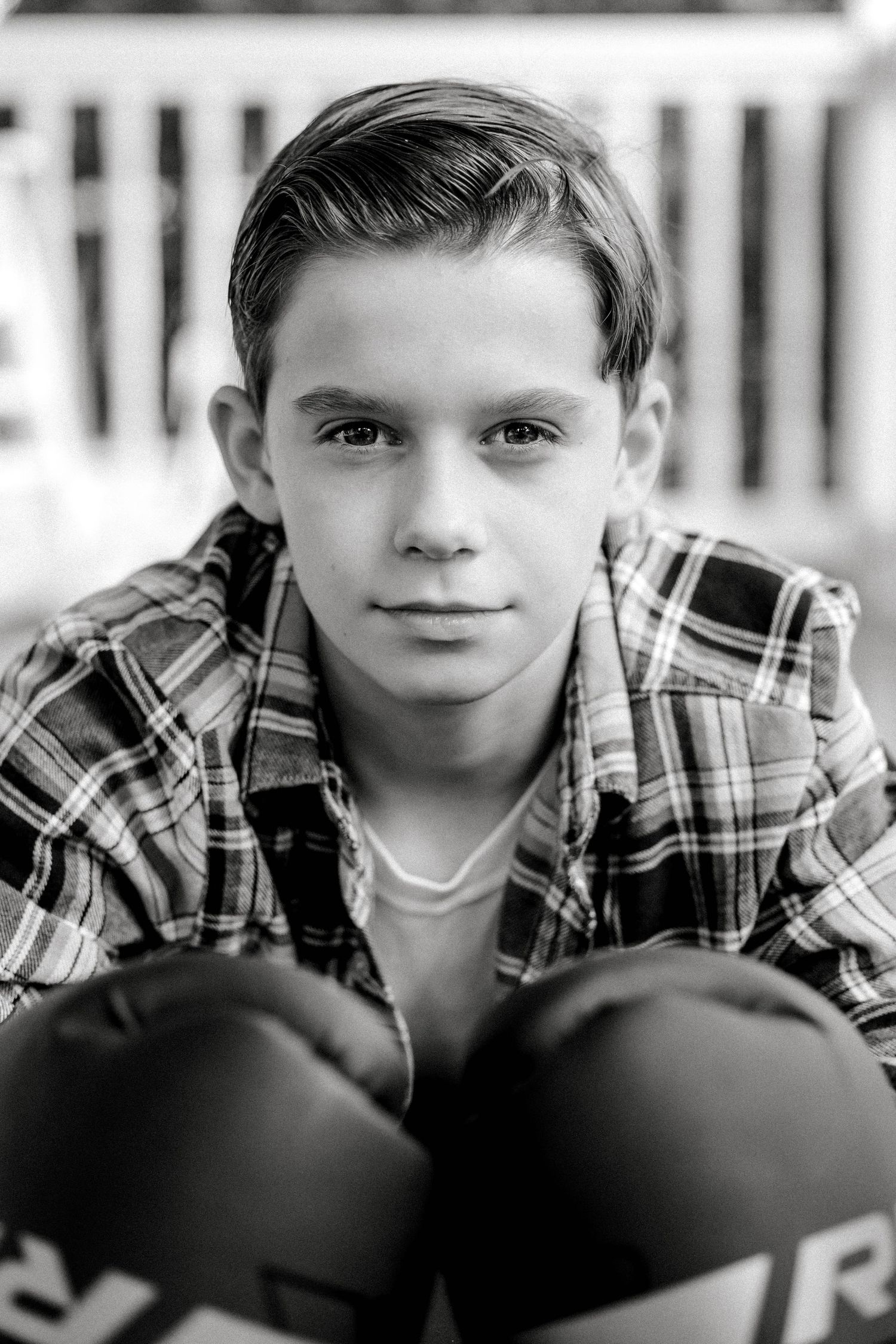 Black and white portrait of a young boy in a plaid shirt wearing boxing gloves looking seriously at camera.