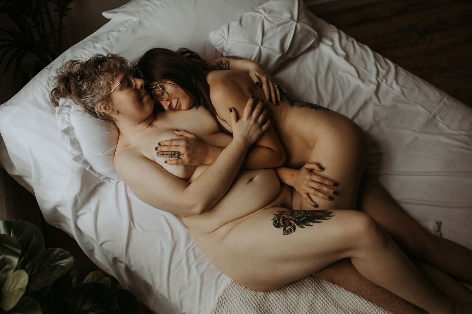 Person wearing black lace lingerie lounges on white bedding in moody boudoir photography setting.