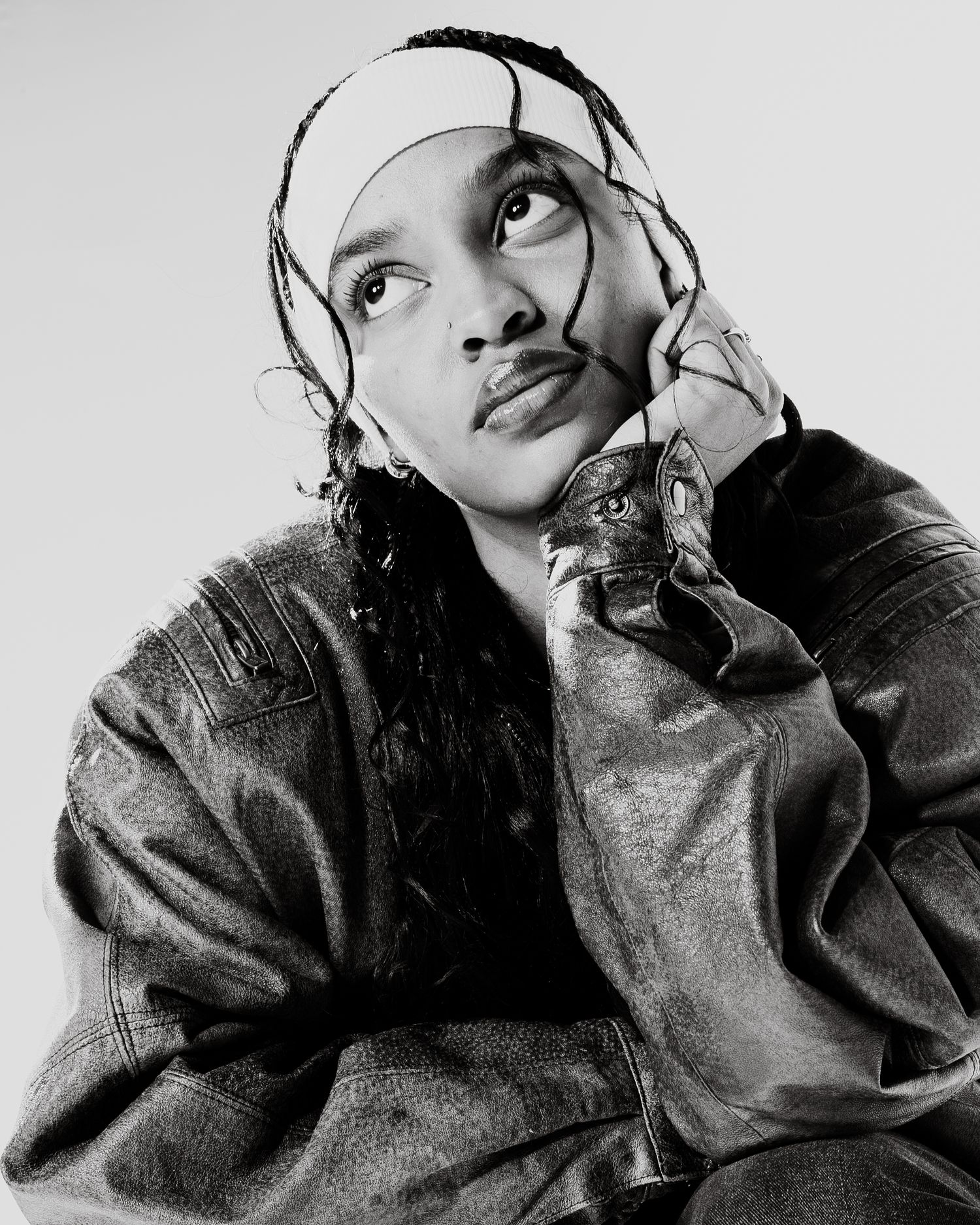 Black and white portrait of a person with braids wearing leather jacket, gazing upward thoughtfully.