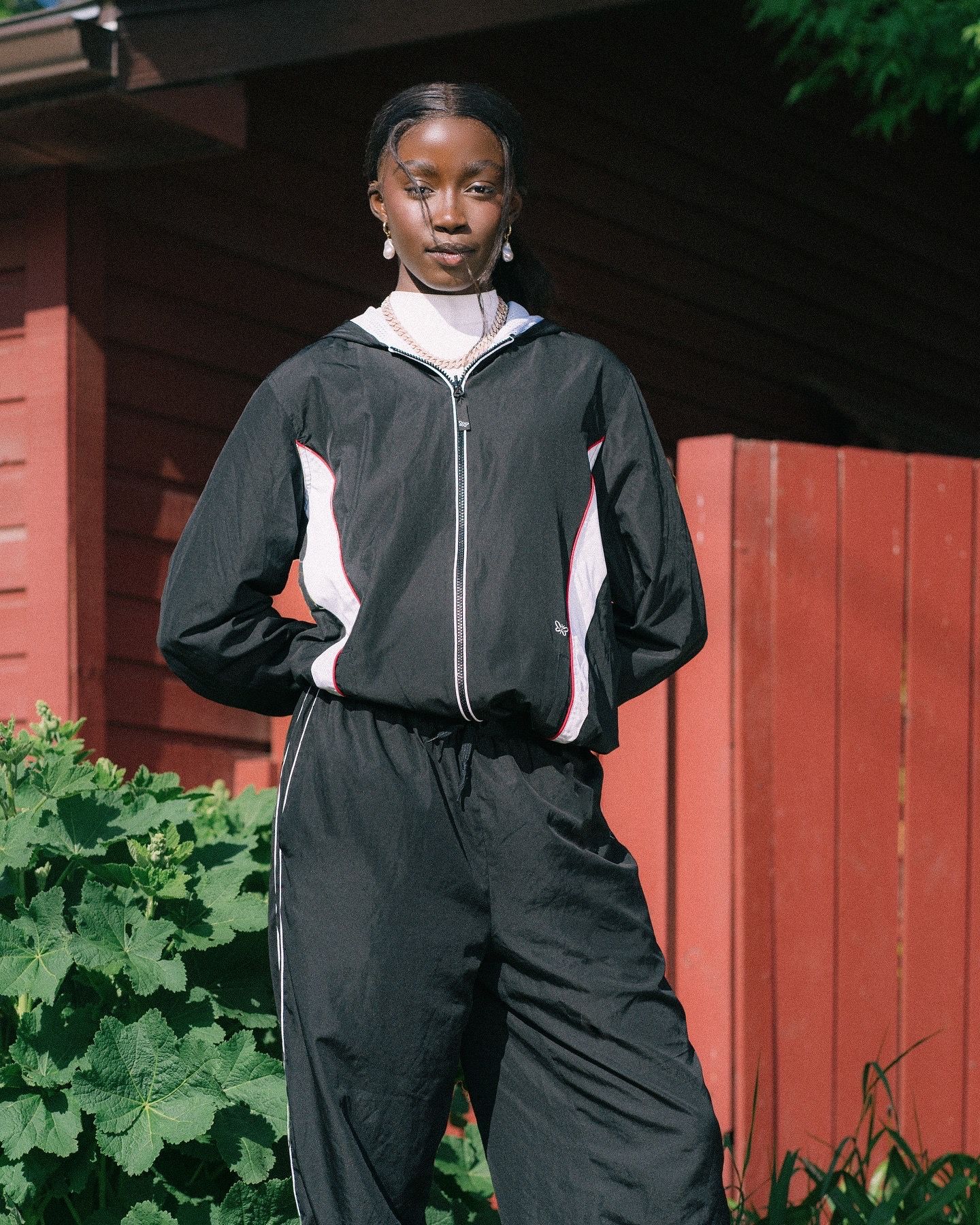 Young Black woman posing outdoors in black and pink windbreaker tracksuit set against red wooden fence.
