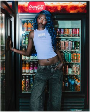 Woman with blue hair wearing crop top and jeans posing in front of illuminated Coca-Cola beverage cooler.