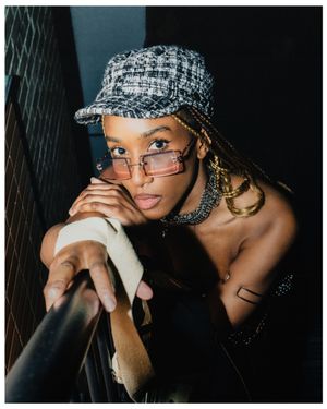 Stylish woman wearing tweed cap, rimless tinted sunglasses, and gold chain necklace posing on dark staircase.