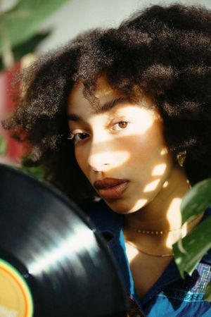 Young woman with natural afro hair holding a vinyl record with dappled sunlight across her face.