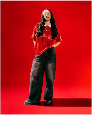 A woman in a red polo shirt and wide-leg black jeans poses against a bold red background in a streetwear look.
