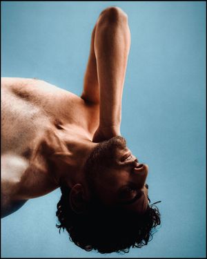 Shirtless man photographed from below against blue background with arm raised, head tilted back.