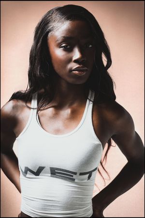 A fit Black woman in a white WET crop tank top poses confidently with hands on hips against a beige background.