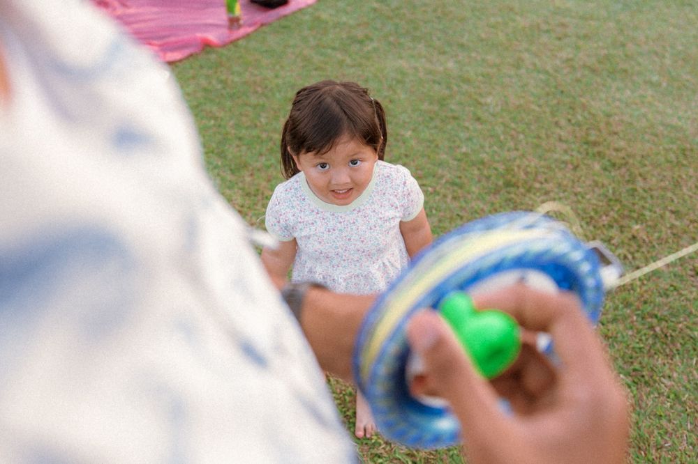 A cheerful toddler in a white dress plays in the grass while someone holds a toy.