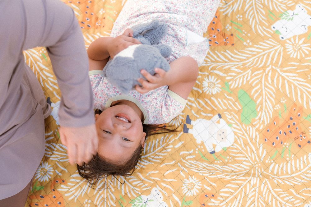 A small child plays and laughs while lying on an orange patterned blanket outdoors.