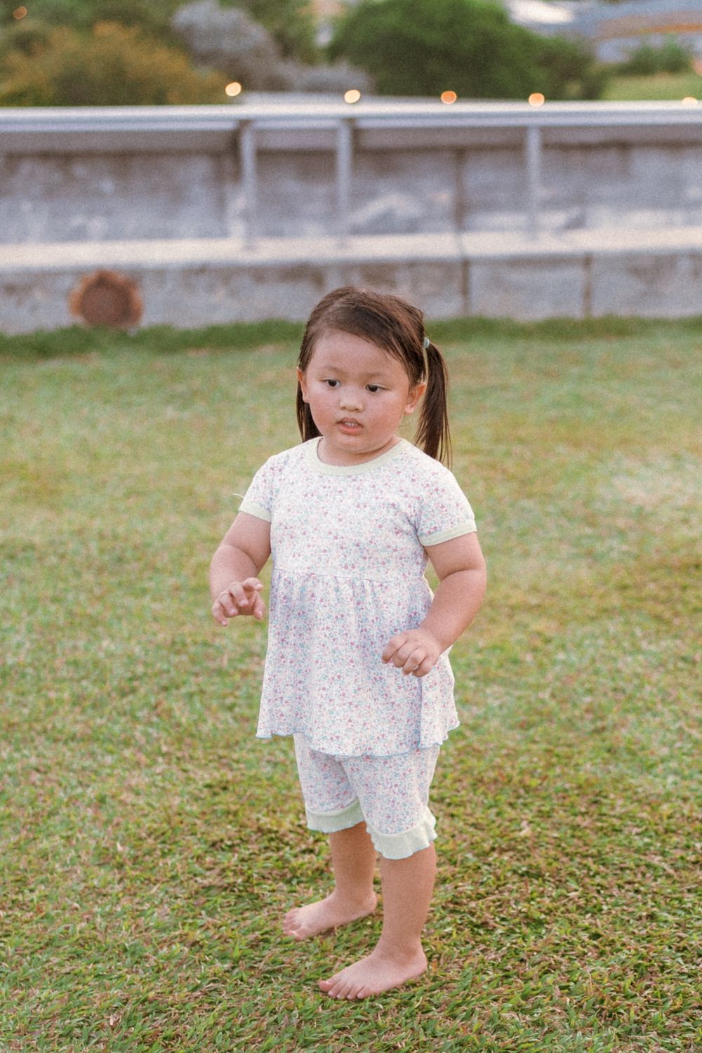 A toddler wearing a white dress and leggings stands barefoot on grass during golden hour.