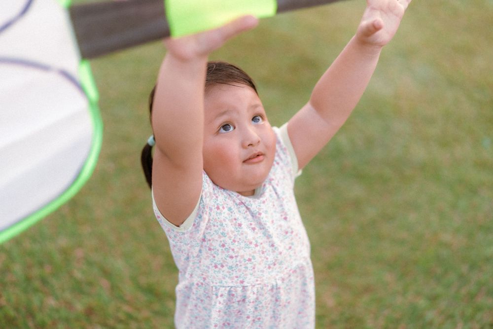 A toddler in a white floral dress raises their arms joyfully while playing outdoors on grass.