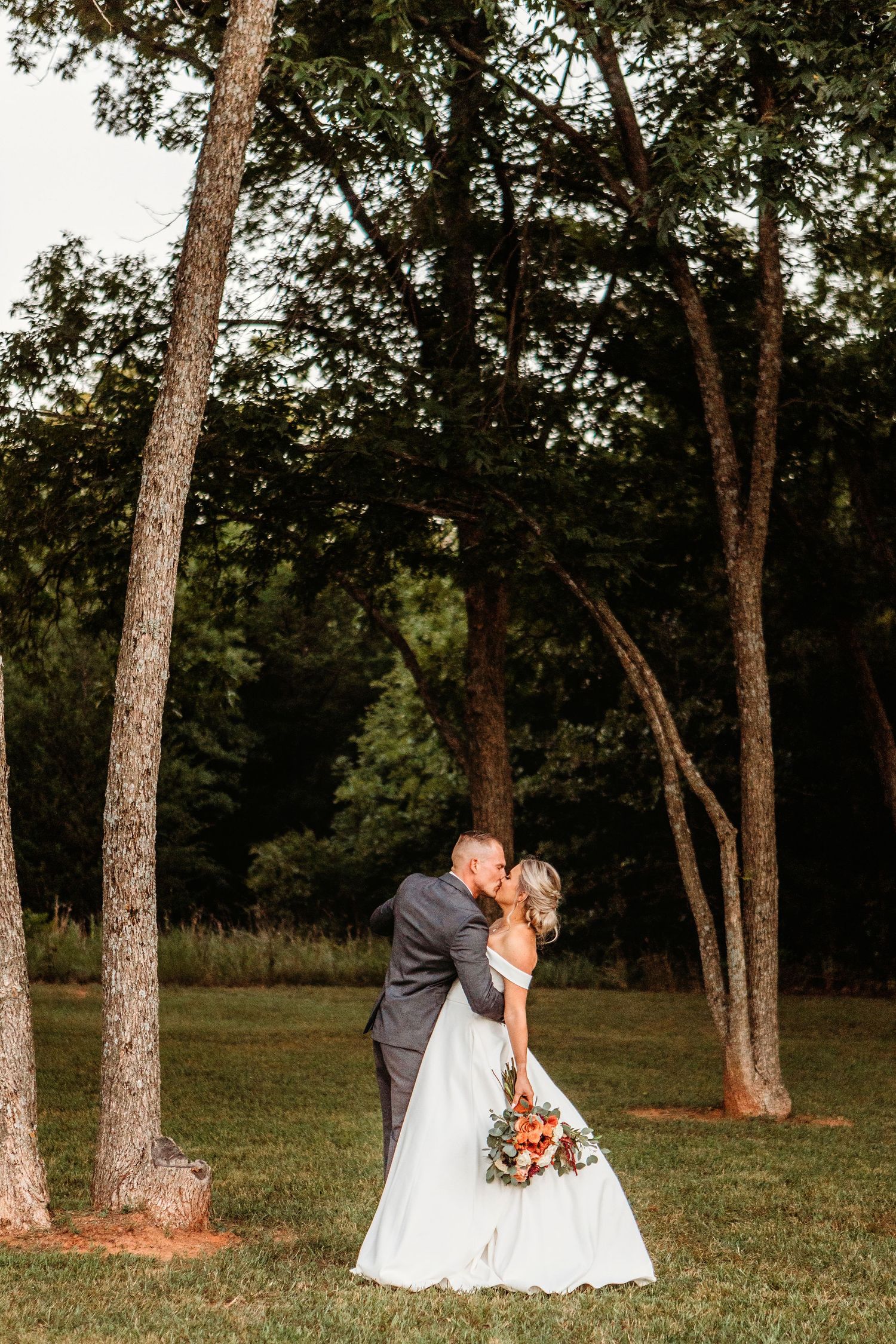 Romantic couple in wedding attire embracing under tall trees in a wooded setting.