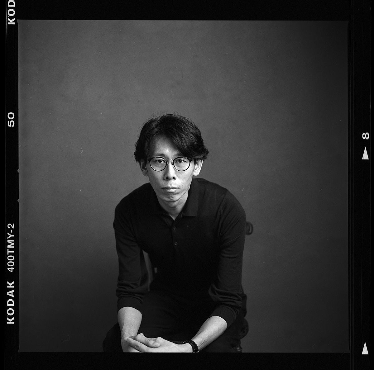 Black and white studio portrait of a person in glasses and dark sweater against simple background. Photographer Noritoshi Ichikawa. Toronto