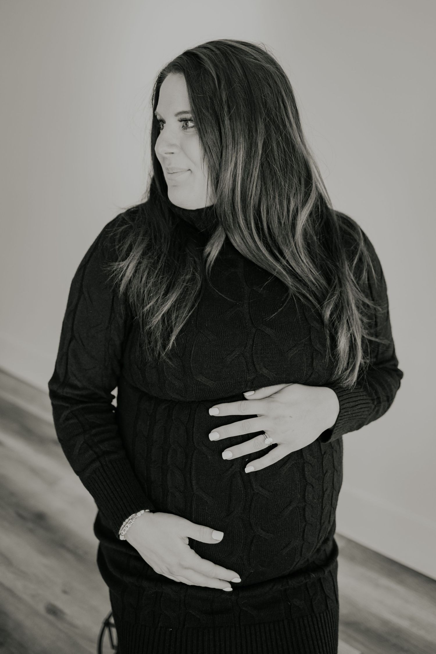 Black and white portrait of a pregnant person wearing a dark sweater while touching their belly in a tender pose.