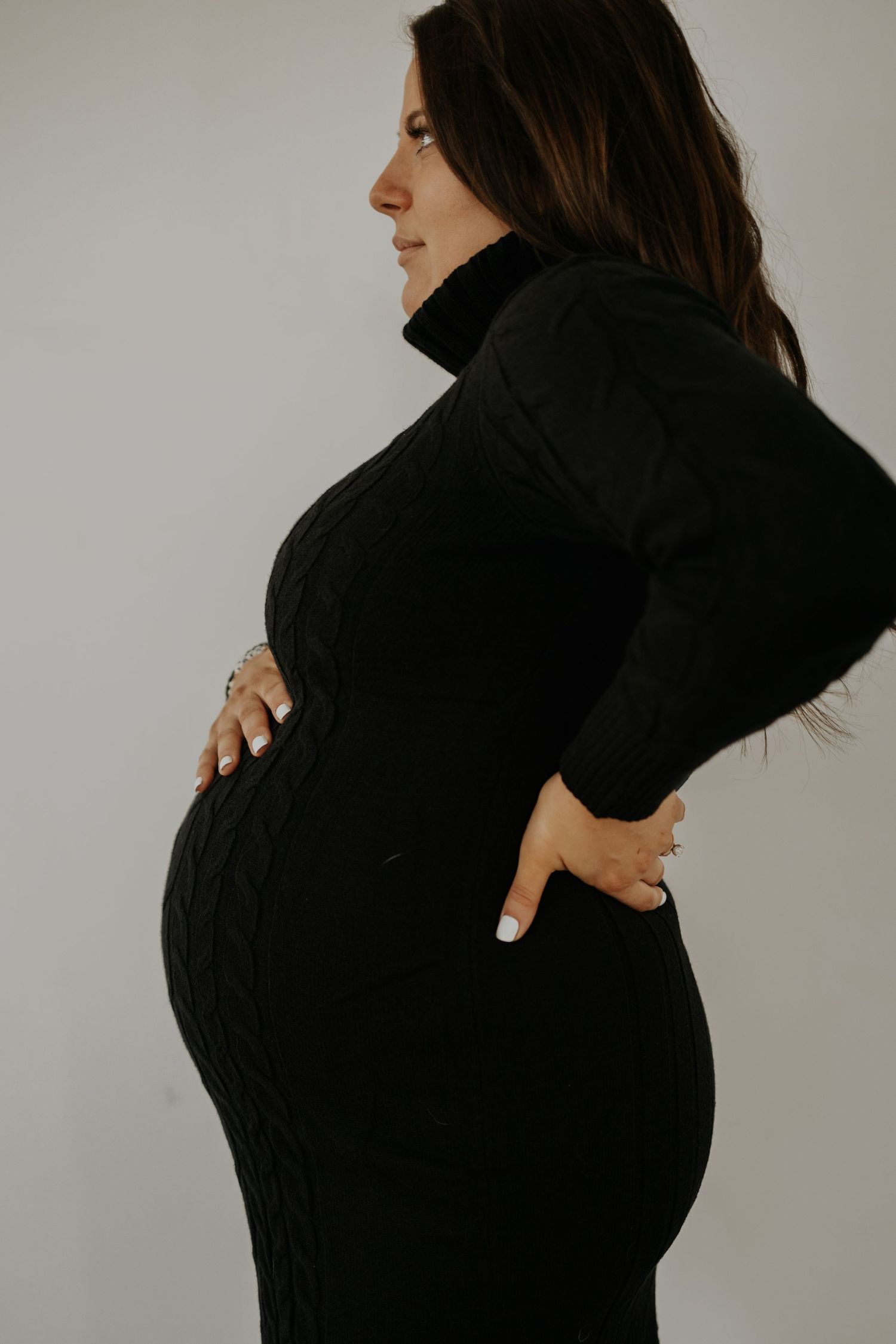 Side profile of expectant mother in black sweater dress holding her belly against neutral background.
