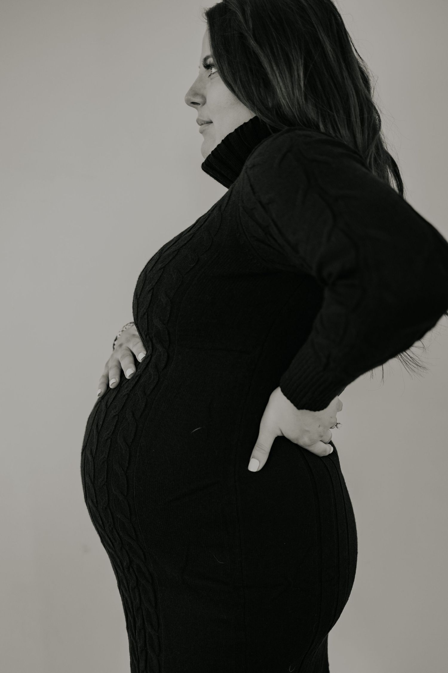 Black and white maternity photo showing silhouette of pregnant woman in dark sweater dress against light background.