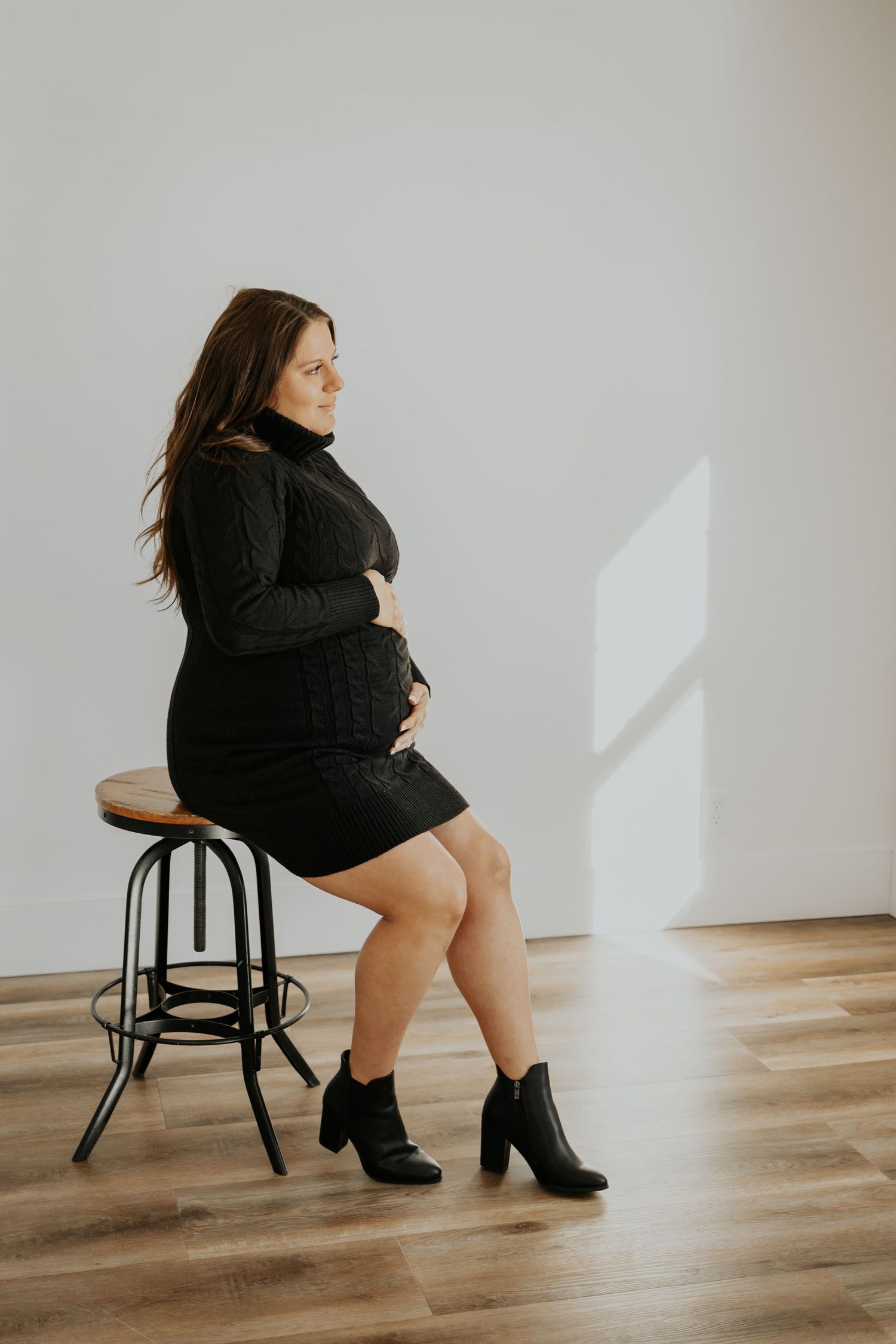 Woman in black dress sits gracefully on bar stool against white wall with sunlight casting geometric shadows.
