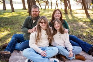 Family relaxing together on blanket under pine trees in park setting.