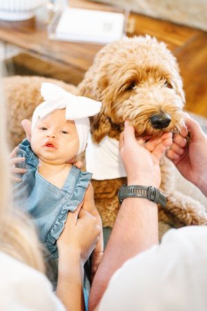 A baby in blue overalls is held and kissed while a fluffy dog watches nearby.