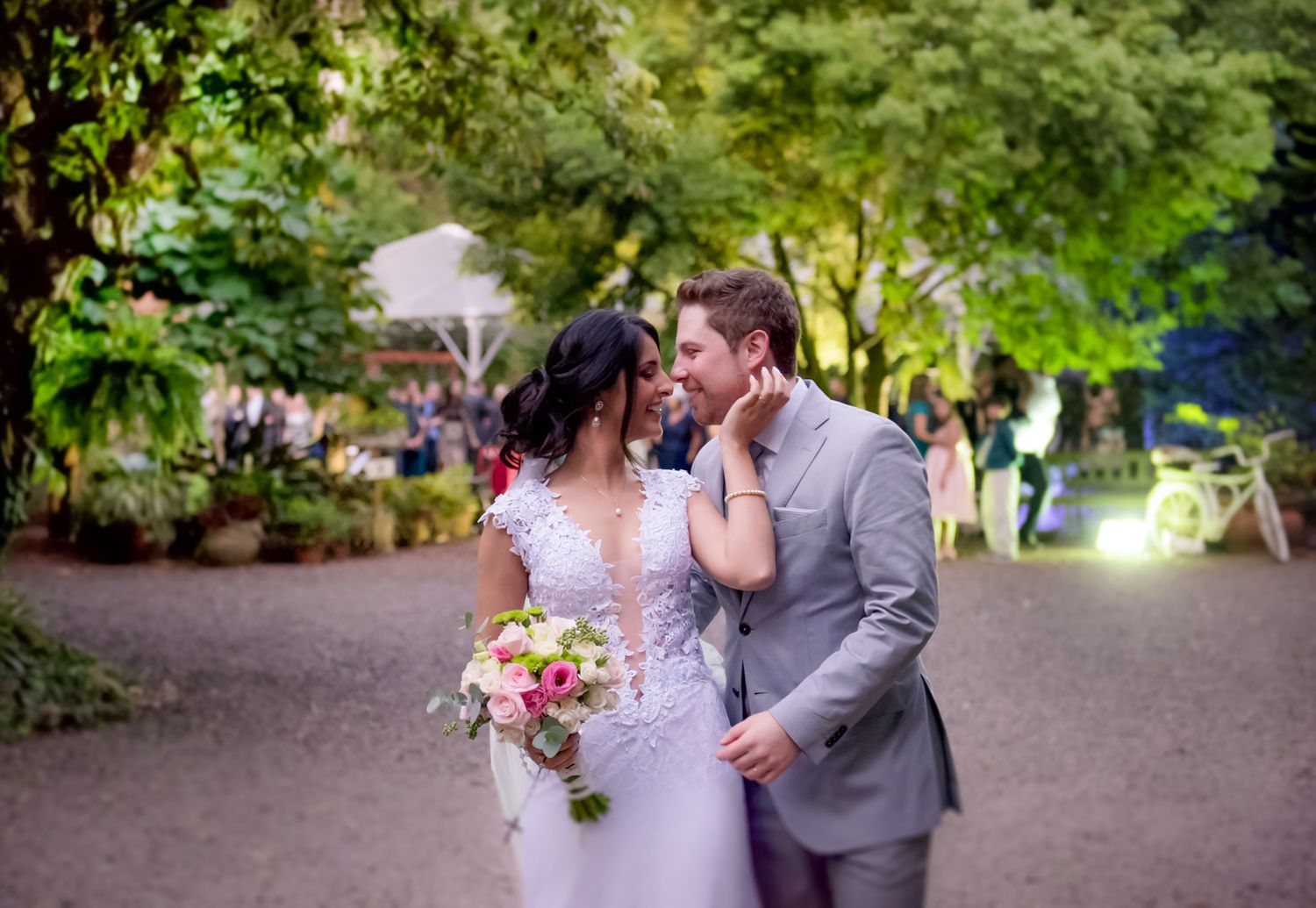 A couple in wedding attire share an intimate moment on a garden path lined with trees and soft lighting.