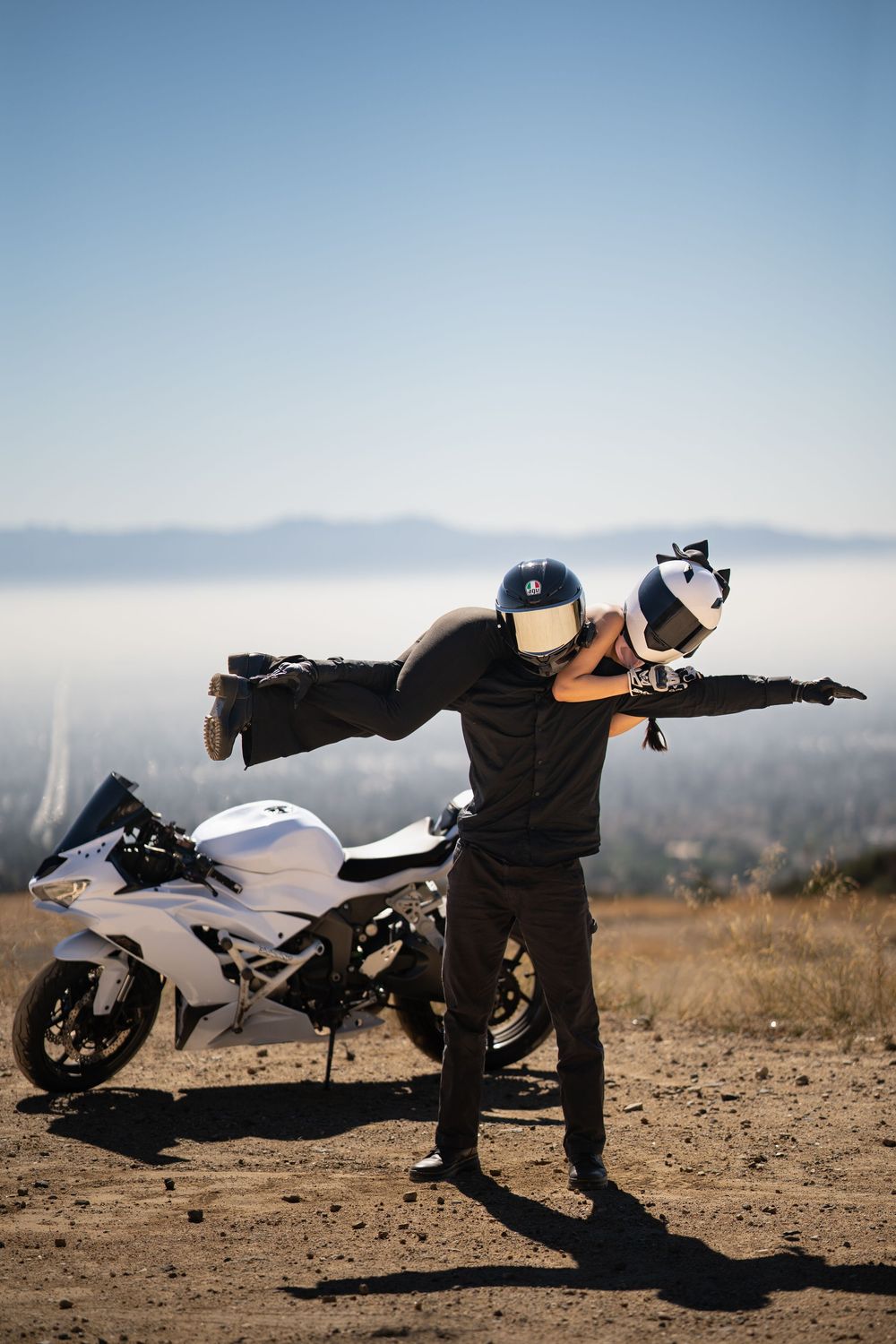 A motorcyclist performs a dynamic yoga pose next to a parked white sport bike against a misty mountain backdrop.