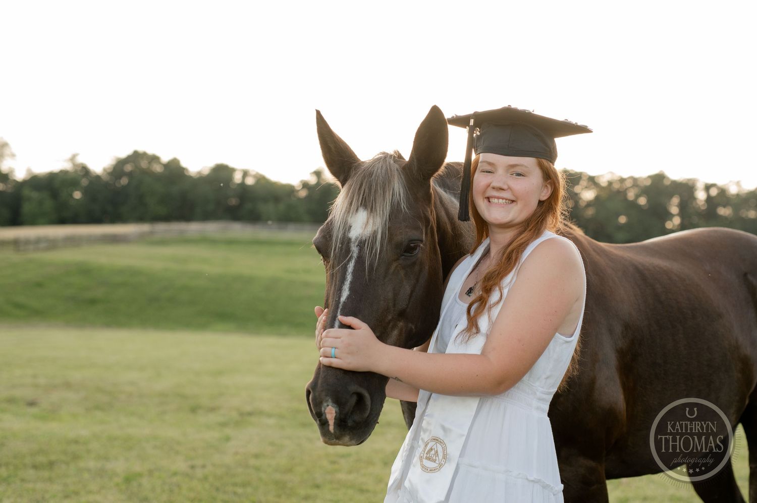 Graduate poses with horse at sunset in pastoral setting wearing cap and gown.