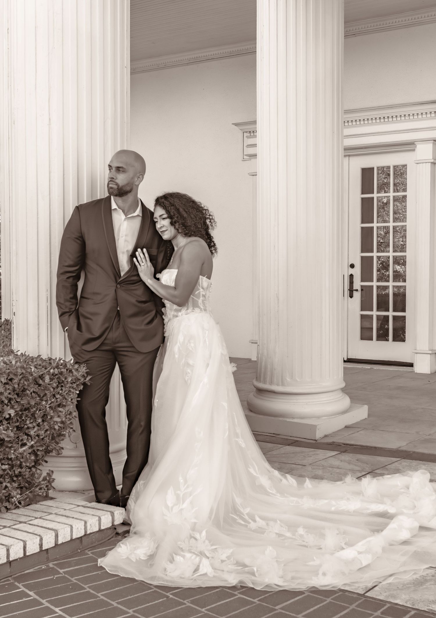 Black and white wedding portrait by elegant white columns and brick pathway.