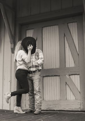 Black and white photo of a couple sharing a kiss, man wearing cowboy hat and boots outside rustic barn doors.