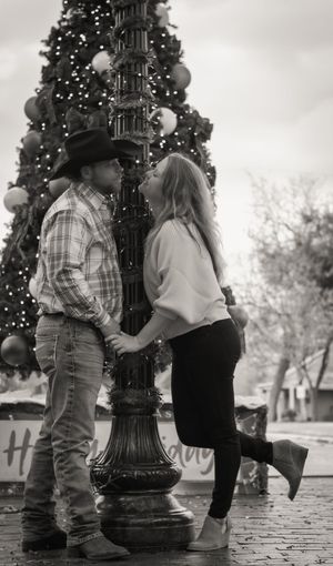 Black and white photo of a couple kissing by a decorated Christmas tree and ornate lamppost outdoors.