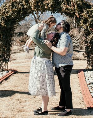 A loving family moment shared outdoors under a floral archway on a sunny day.