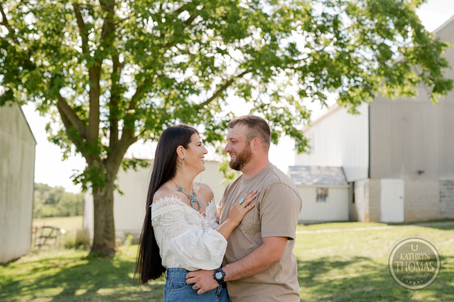 Couple embracing in sunlit outdoor setting with rustic barn and trees.