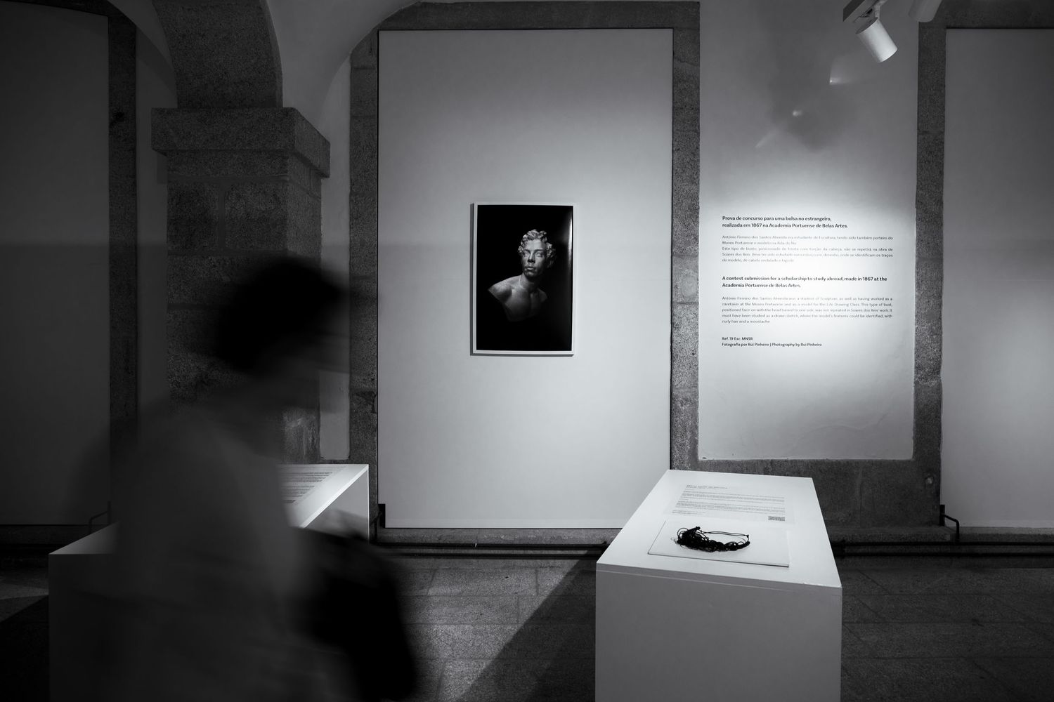 Black and white gallery scene with a framed portrait and blurred visitor movement.