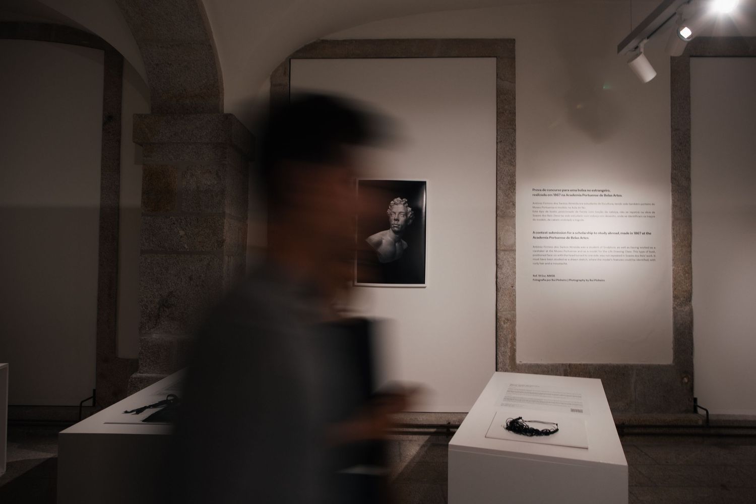 Gallery visitors examine artwork in a dimly lit exhibition space with spotlights illuminating displays.