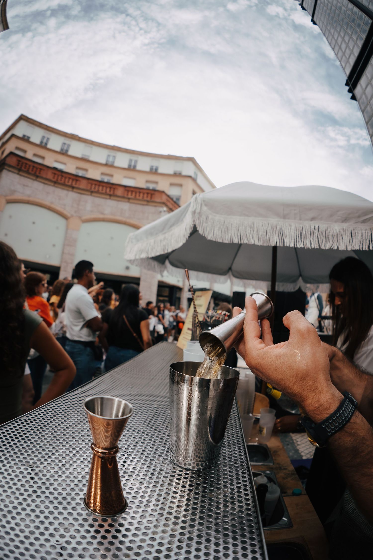 Registo de um bartender a servir num evento ao ar livre com luz suave, toldos brancos e atmosfera descontraída.