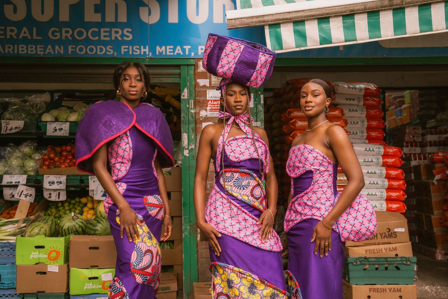 Three women in vibrant African print purple and pink dresses pose outside a grocery store.