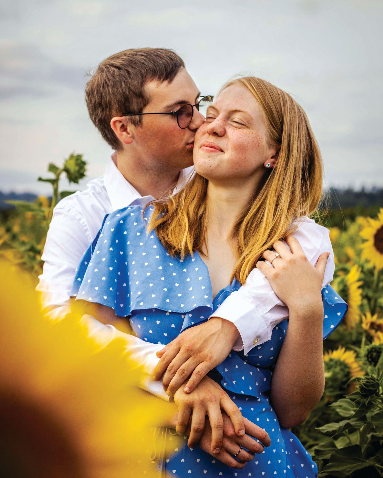 a couple stands in a field of sunflowers, his arms are around her and she smiles as he kisses her cheek