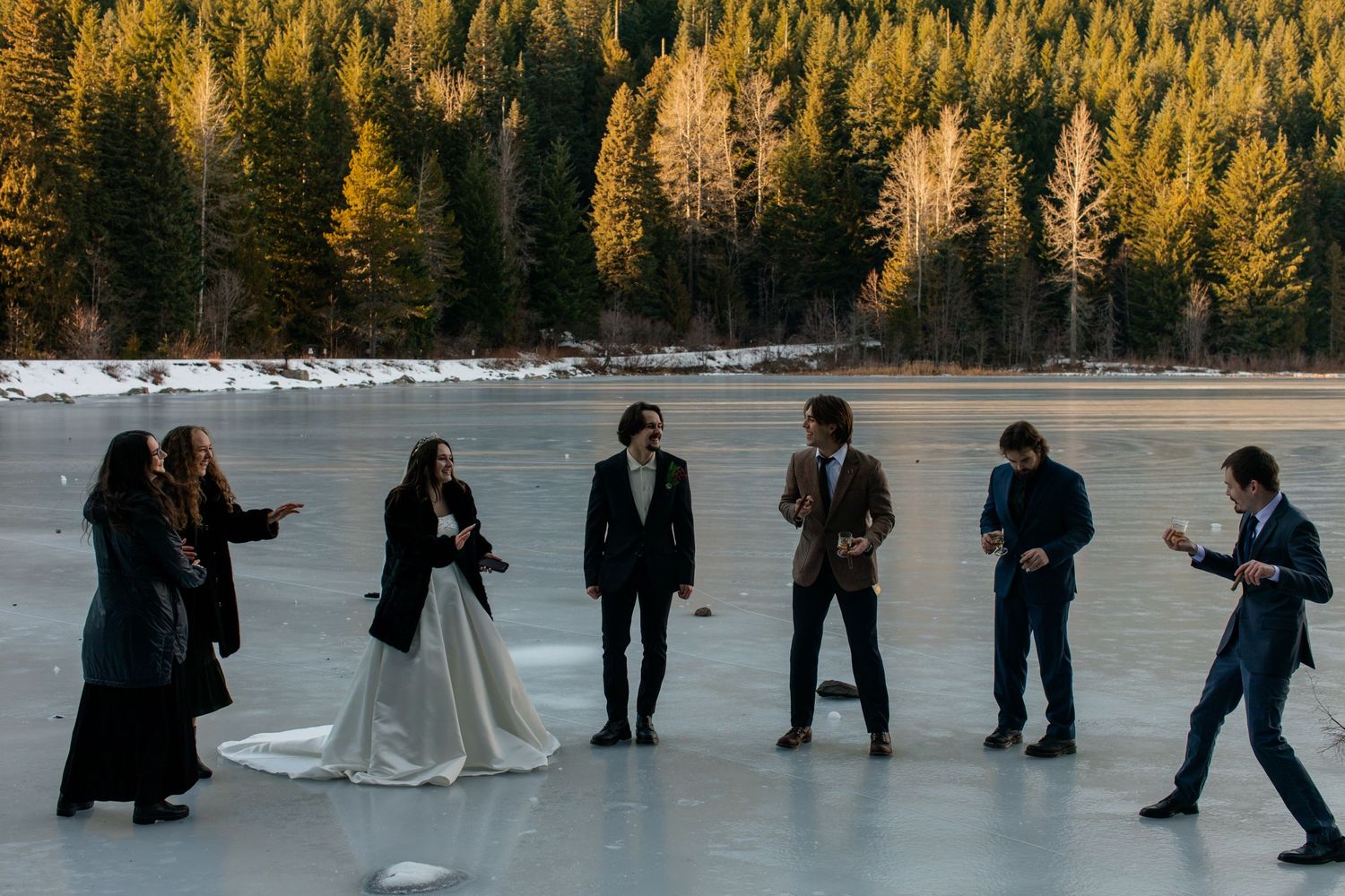 Wedding party celebrating on a frozen lake with evergreen forest backdrop.