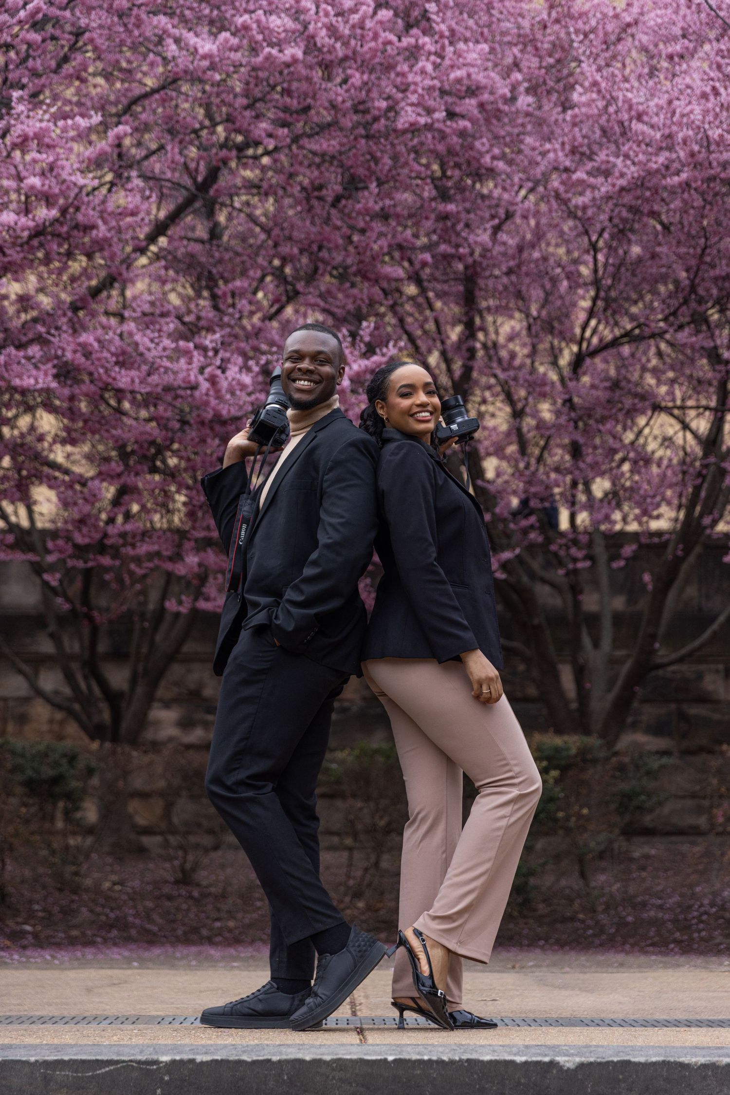 Smiling couple with cameras posing back-to-back in front of pink cherry blossom trees.