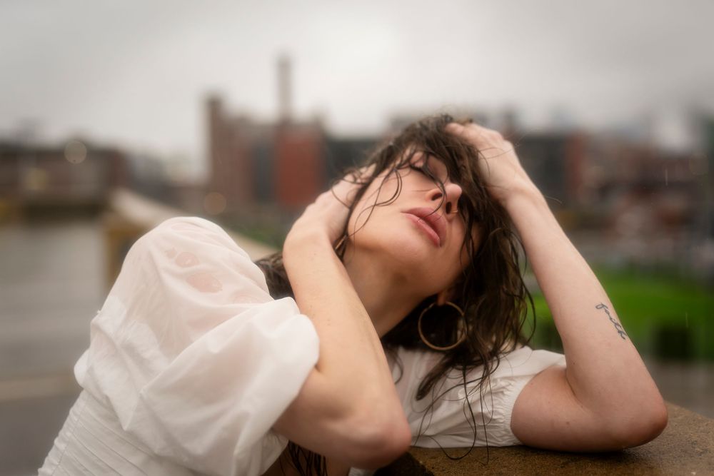 Someone wearing a white dress rests with their head tilted back against a railing near an urban waterfront.