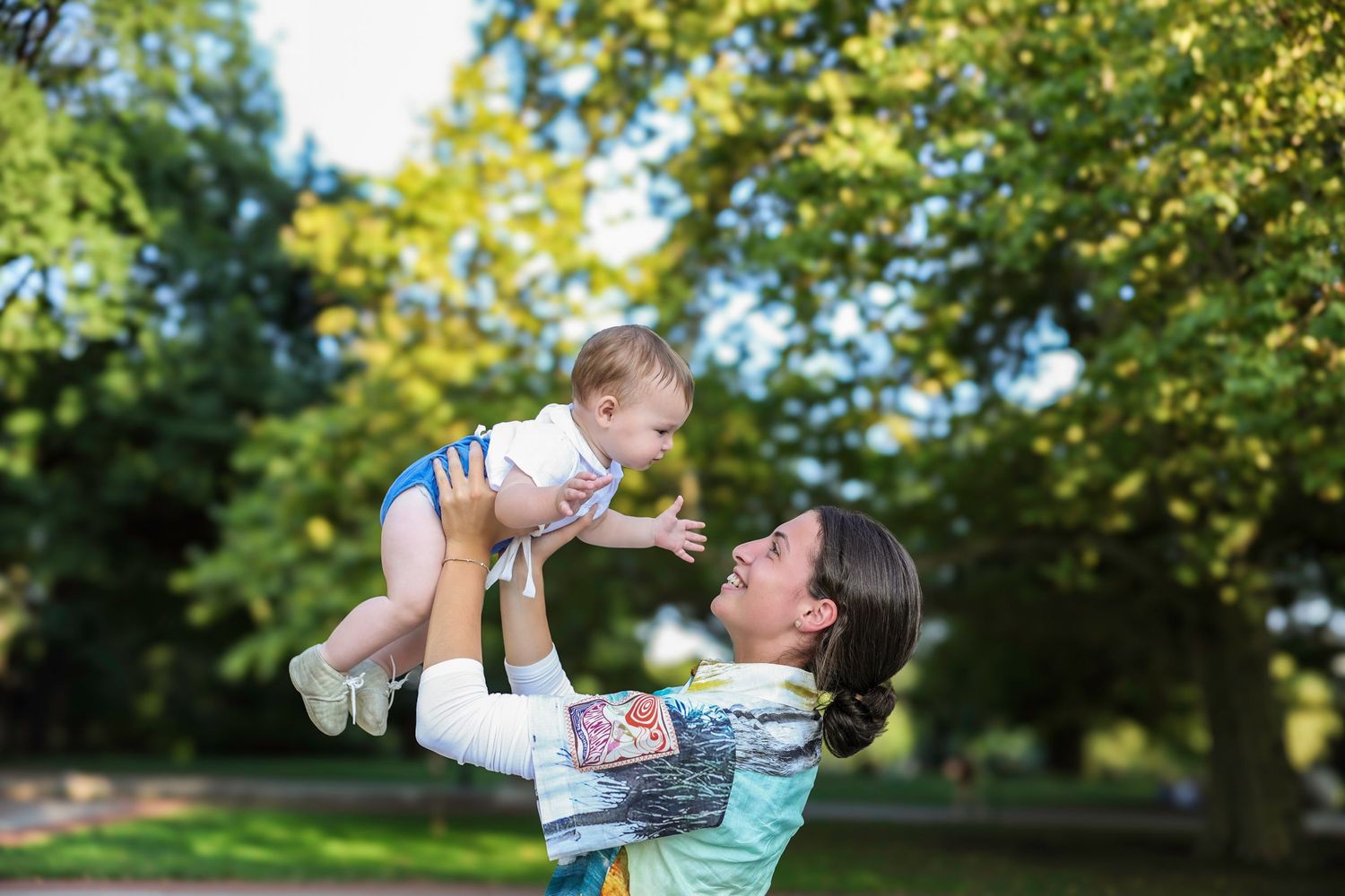 A mother joyfully lifts her laughing baby in the air in a sunny green park.