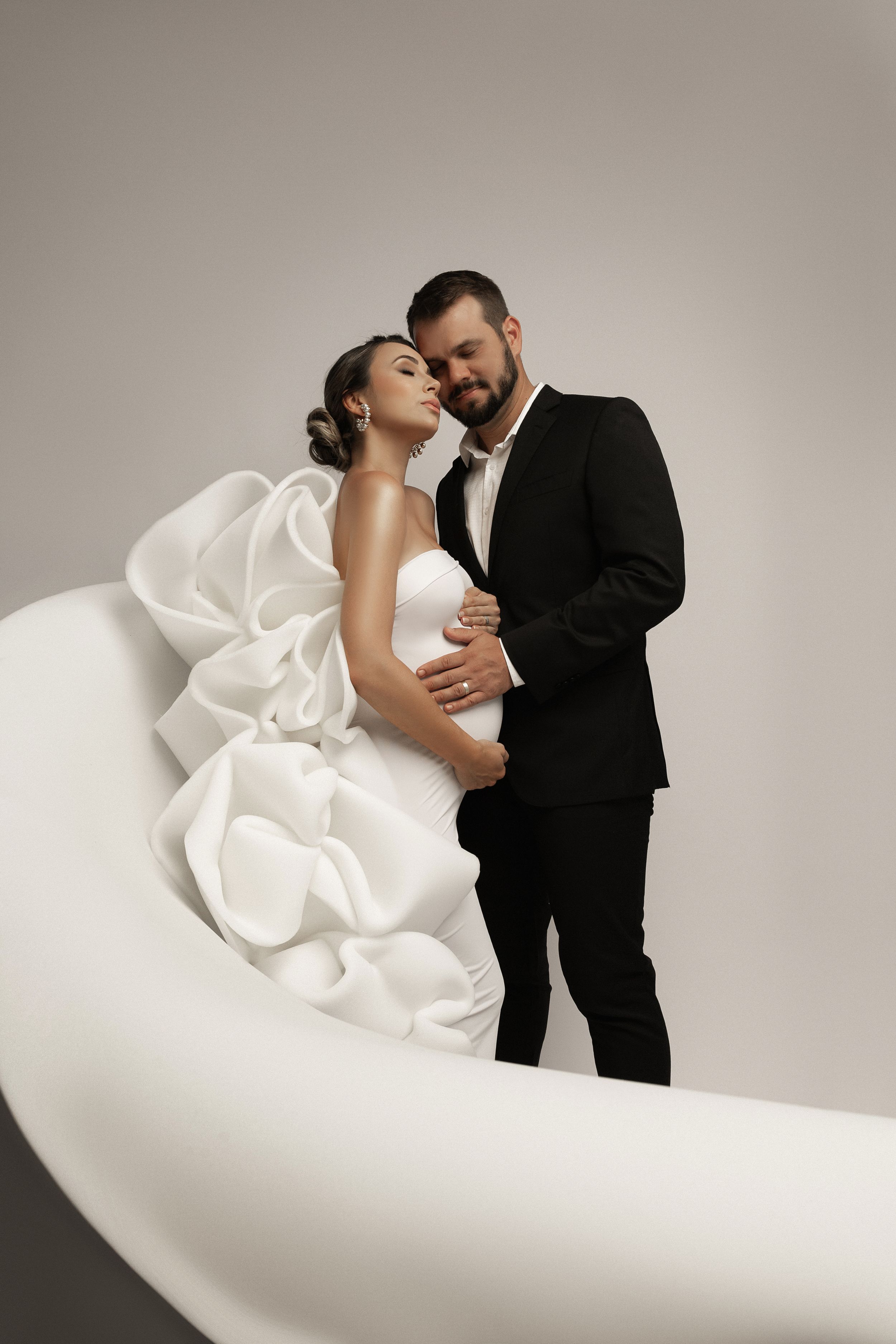 Elegant couple embracing in formal attire against minimalist curved backdrop.