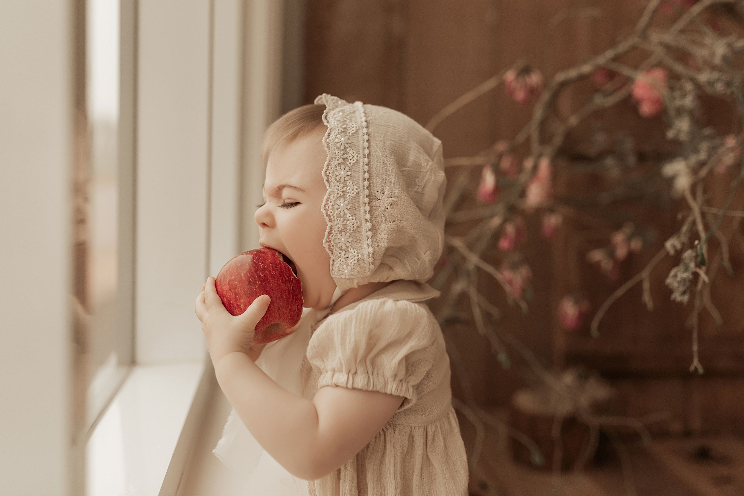 Child in vintage bonnet takes a bite from red apple by window.