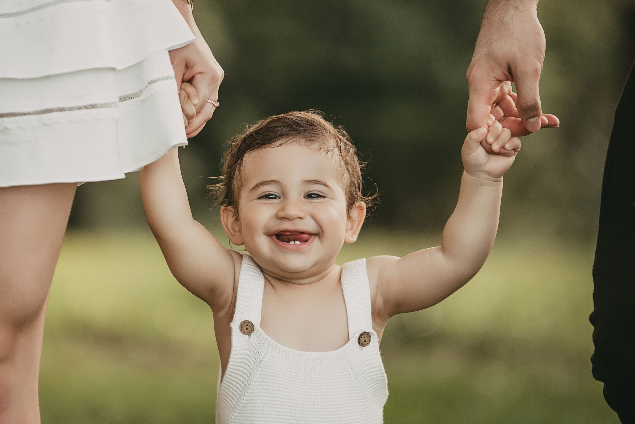 Toddler in white overalls smiles while holding hands during outdoor walk.